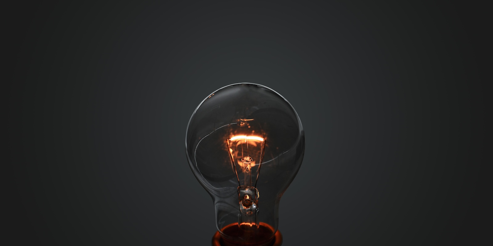 A photo of an incandescent light bulb with lit filament over gray gradient background.